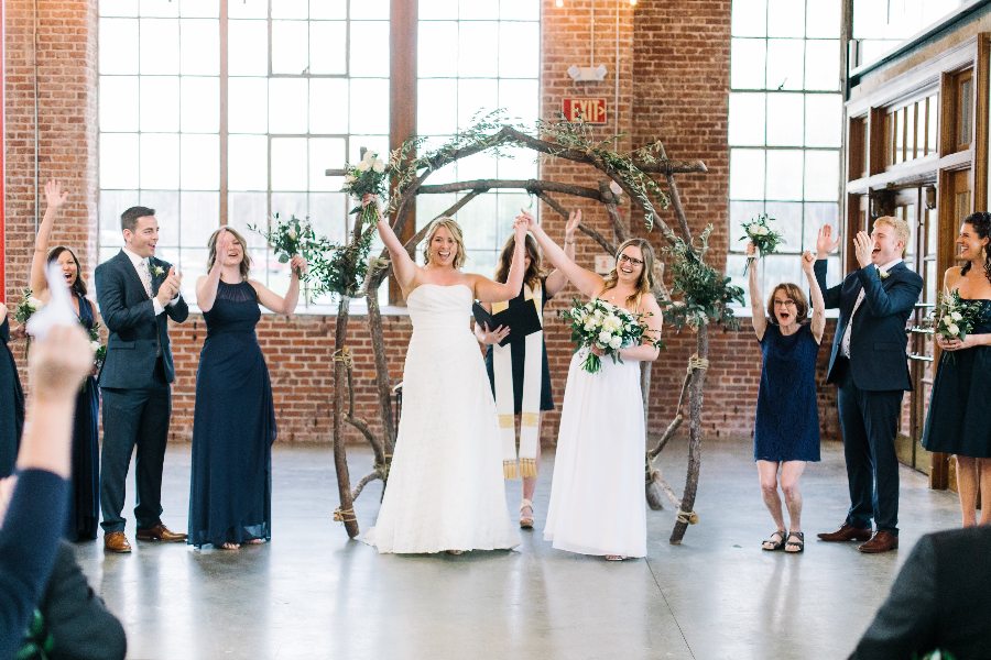 Two brides raising their hands as their wedding party cheers at the end of the ceremony