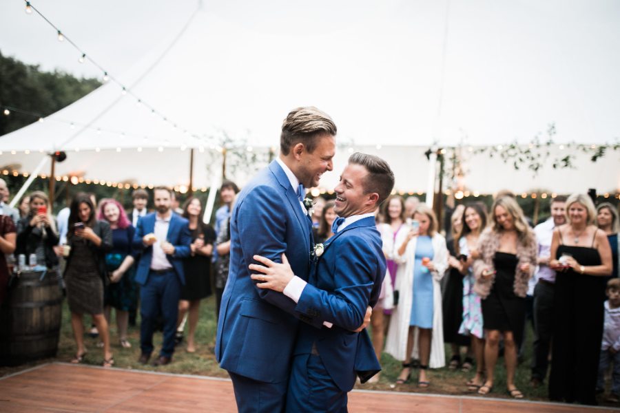 Two grooms in blue suits laughing during their first dance under a tent